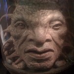 Huge disembodied face with scales and tentacles in a jar.