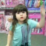 young girl with bangs stands in front of dolls in pink boxes